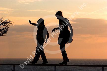 Silhouette of children walking on a railing at sunset by the sea.
