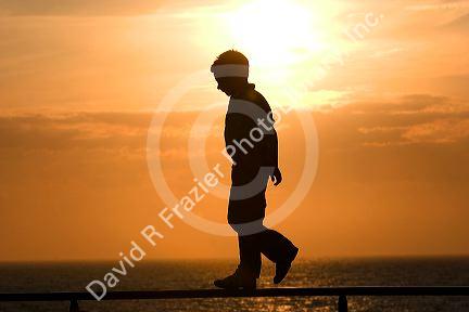 Silhouette of a child walking on a railing at sunset by the sea.