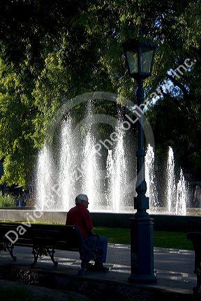 Man sitting on a park bench in front of a water fountain in Plaza Independencia in Mendoza, Argentina.