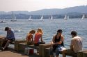 People sit along the Zurichsee Lake with sailboats behind at Zurich, Switzerland.

switzerland, swiss, europe, european, travel, tourism, swiss alps, alps, alpine, zurich, sailboat, sail boat, boat, zurichsee lake, zurichsee, lake