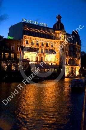 NH Doelen Hotel with lights on at night along the Amstel River in Amsterdam, Netherlands.