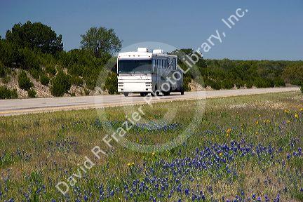 A motorhome recreational vehicle pulling a trailer on Interstate 10 in west Texas with Texas blue bonnet wildflowers in the median.